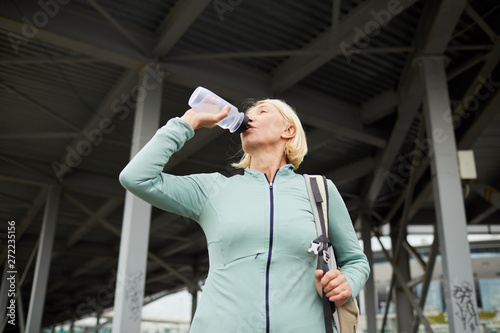 Blonde mature thirsty sportswoman drinking water from plastic bottle after hard training in urban environment on summer day