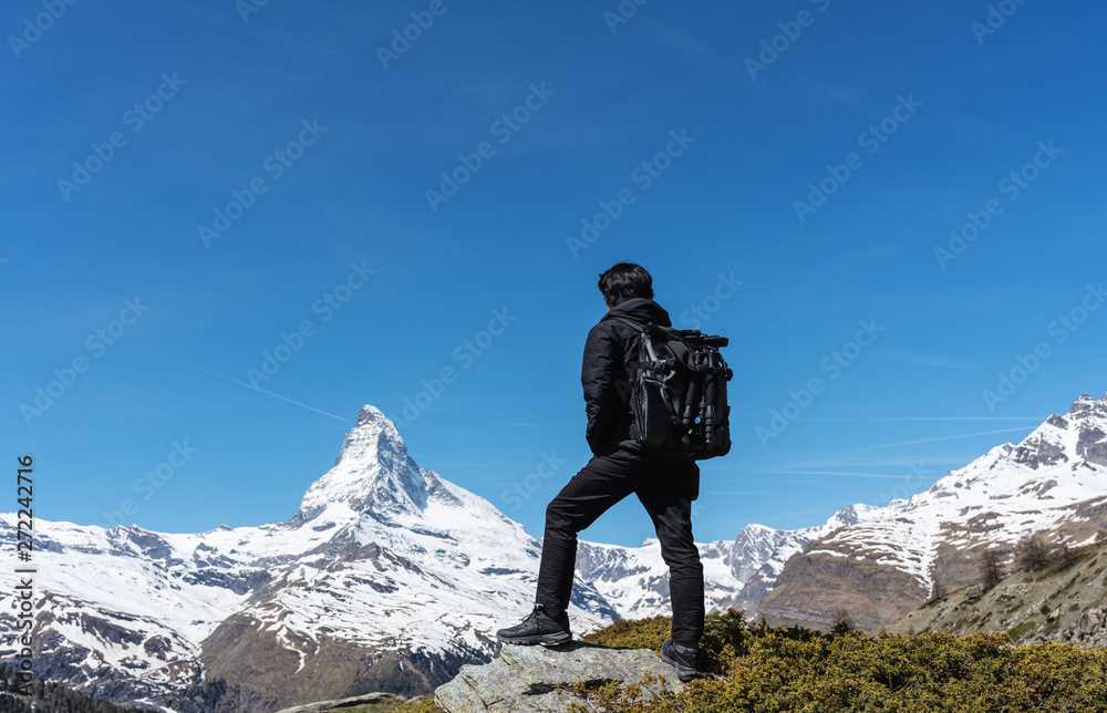 Travelling in Switzerland, a man with backpack standing on the rock looking at Matterhorn mountain view. Trekking in Zermatt, Switzerland Travelling in Switzerland, a man with backpack standing on the rock looking at Matterhorn mountain view. Trekking in Zermatt, Switzerland