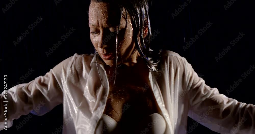 portrait of a dark-Haired girl in a white shirt and a white bra, she stands on a black background in the rain, dancing.