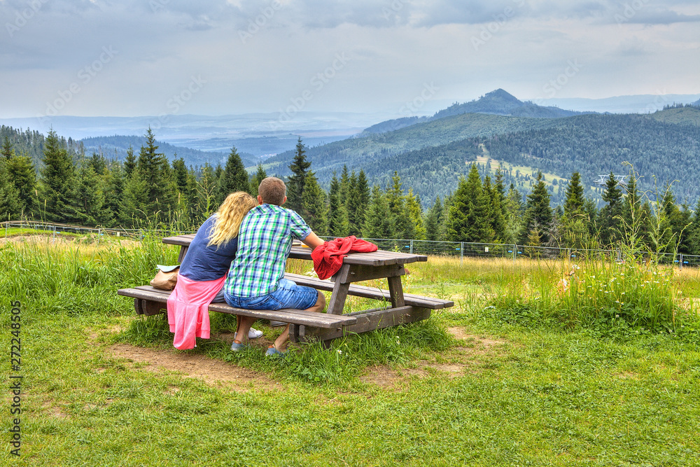 Romantic couple sitting on the wooden bench at the top of the hill and overlooking the view on mountain peaks. Young man and woman in love sitting together and watching beautiful scenery of mountains. Romantic couple sitting on the wooden bench at the top of the hill and overlooking the view on mountain peaks. Young man and woman in love sitting together and watching beautiful scenery of mountains.
