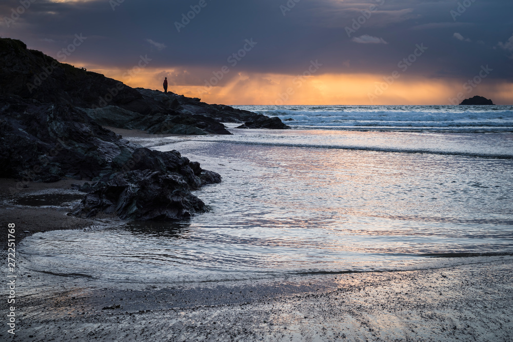A distant figure looking out to sea and silhouetted against the dramatic sky at sunset. Polzeath, Cornwall UK.