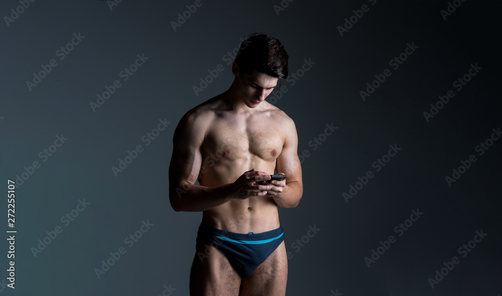 Low key portrait of muscular shirtless man at dark background standing with smart phone