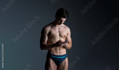 Low key portrait of muscular shirtless man at dark background standing with smart phone