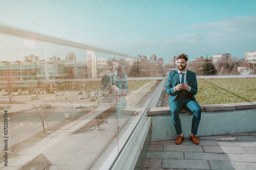 Young smiling white collar worker in blue suit sitting on the rooftop with headphones on ears and using smart phone while looking at beautiful view.