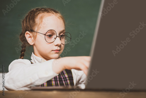 Little student sitting at table and writing on notebook. Kid with a book. Child is ready to answer with a blackboard on a background. Close up.