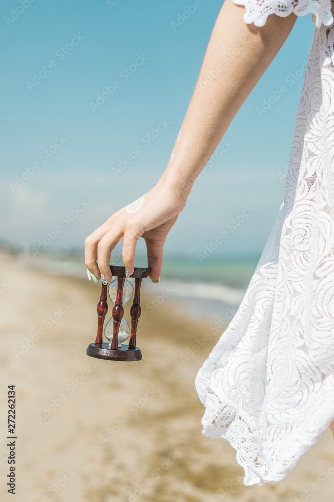 Fototapeta premium Hourglass in female hand on the seashore. Time to rest, relax concept.