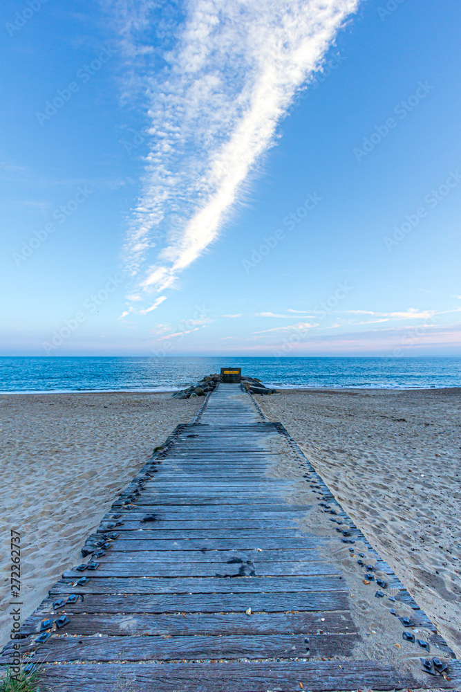 A view of a wooden ponton on a sandy beach with stony groyne (breakwater) and calm beautiful sea in the background at blue hour under a majestic blue sky and white clouds