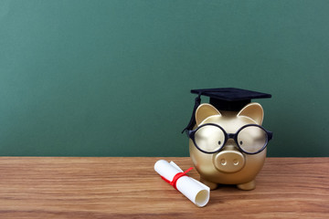 Gfold piggy bank with a grad cap and diploma in front of green chalkboard. Education scholarship