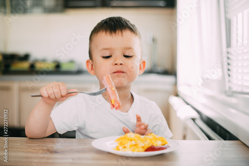 a child in a t-shirt in the kitchen eating a sausage and an omelet with a fork is very appetizing