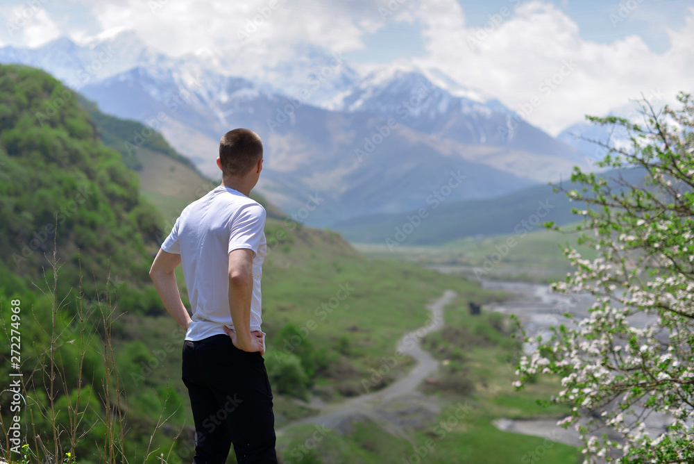 The young man stands high on the mountain next to a flowering tree and looks at the valley and snow-capped mountains in the warm Sunny spring.
