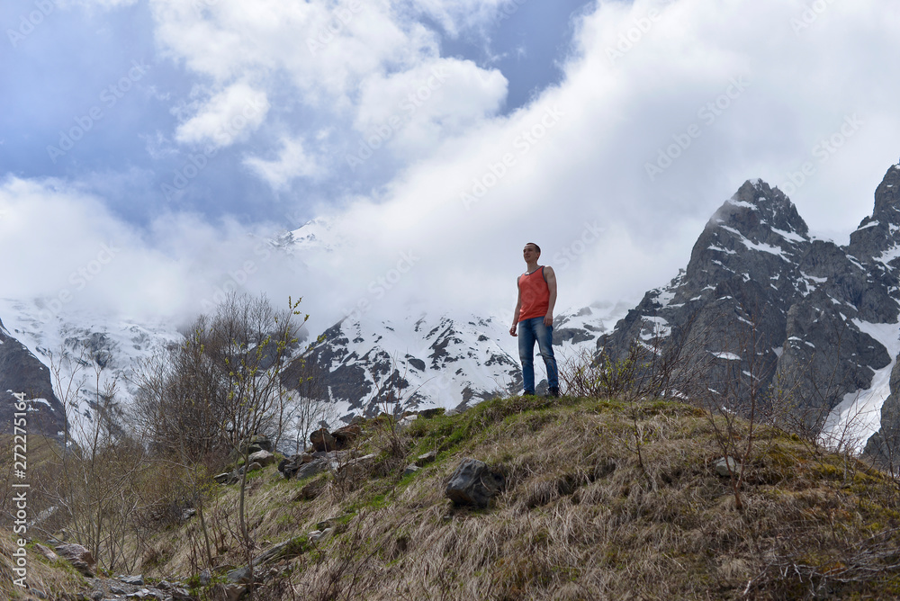 Athletic young man in the orange shirt on top of snow and the rocky mountains on a warm Sunny spring day.