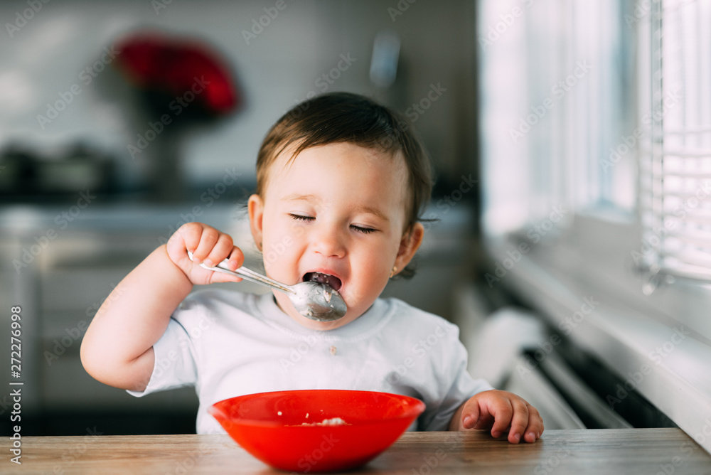 Funny little girl in the kitchen during the day eating oatmeal porridge from a red plate all dirty Funny little girl in the kitchen during the day eating oatmeal porridge from a red plate all dirty