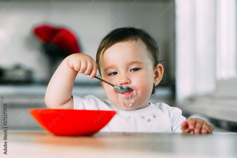 Funny little girl in the kitchen during the day eating oatmeal porridge from a red plate all dirty Funny little girl in the kitchen during the day eating oatmeal porridge from a red plate all dirty