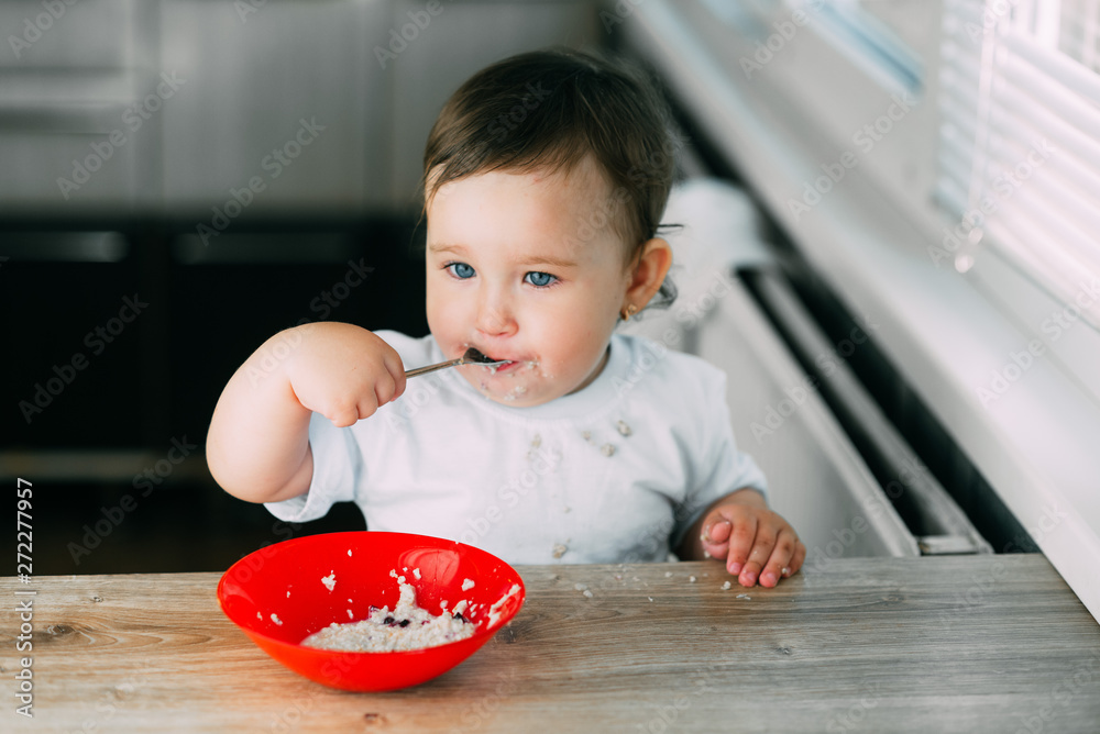 Funny little girl in the kitchen during the day eating oatmeal porridge from a red plate all dirty Funny little girl in the kitchen during the day eating oatmeal porridge from a red plate all dirty