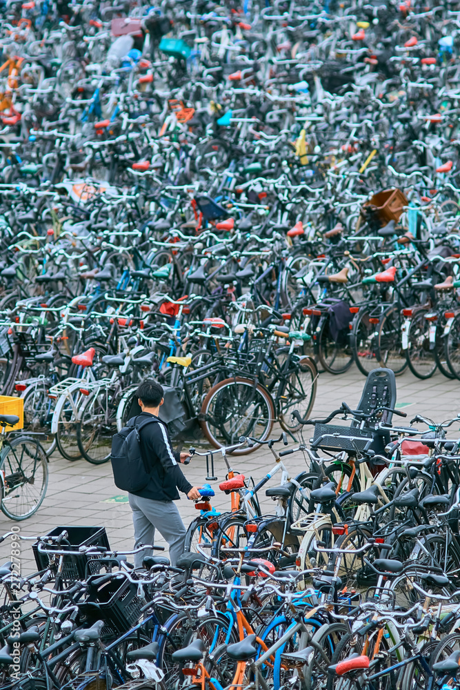 Young asian man searching for a parking space for his bicycle being lost in an overcrowded parking lot for bicycles in Amsterdam, Netherlands.