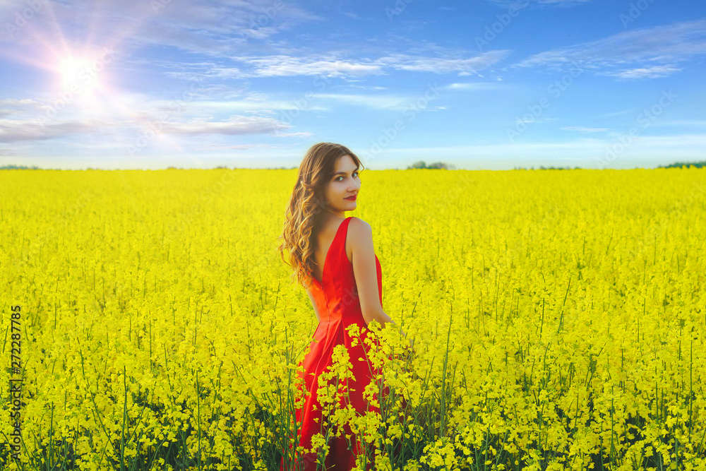 Young beautiful girl in a red dress close up in the middle of yellow field with radish flowers and sunlight..