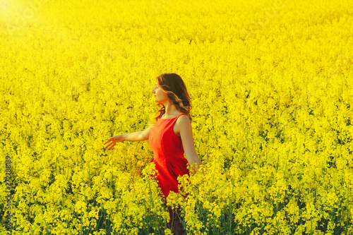 Young beautiful girl in a red dress close up in the middle of yellow field with radish flowers and sunlight.. © volhavasilevich