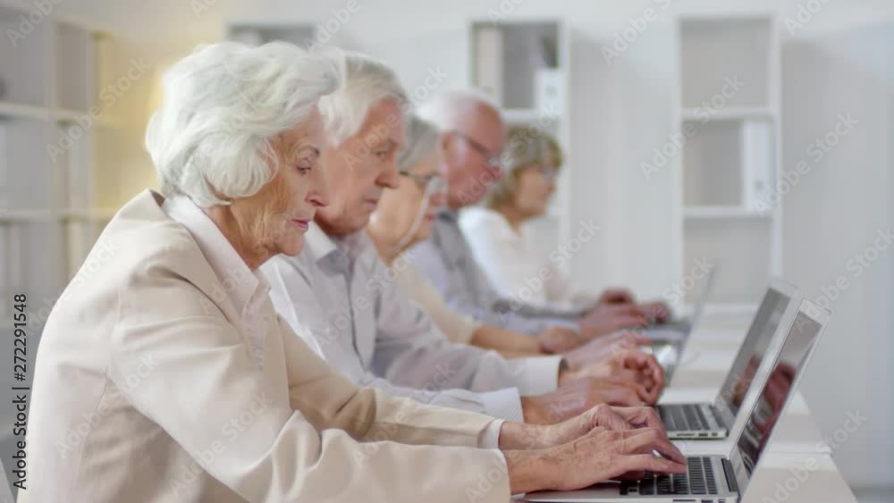 Tracking shot of group of elderly men and women with grey hair sitting at desks and typing on laptops in computer class Tracking shot of group of elderly men and women with grey hair sitting at desks and typing on laptops in computer class