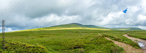 A panoramic view of a mountain with grassy green slope under a majestic blue sky and white clouds