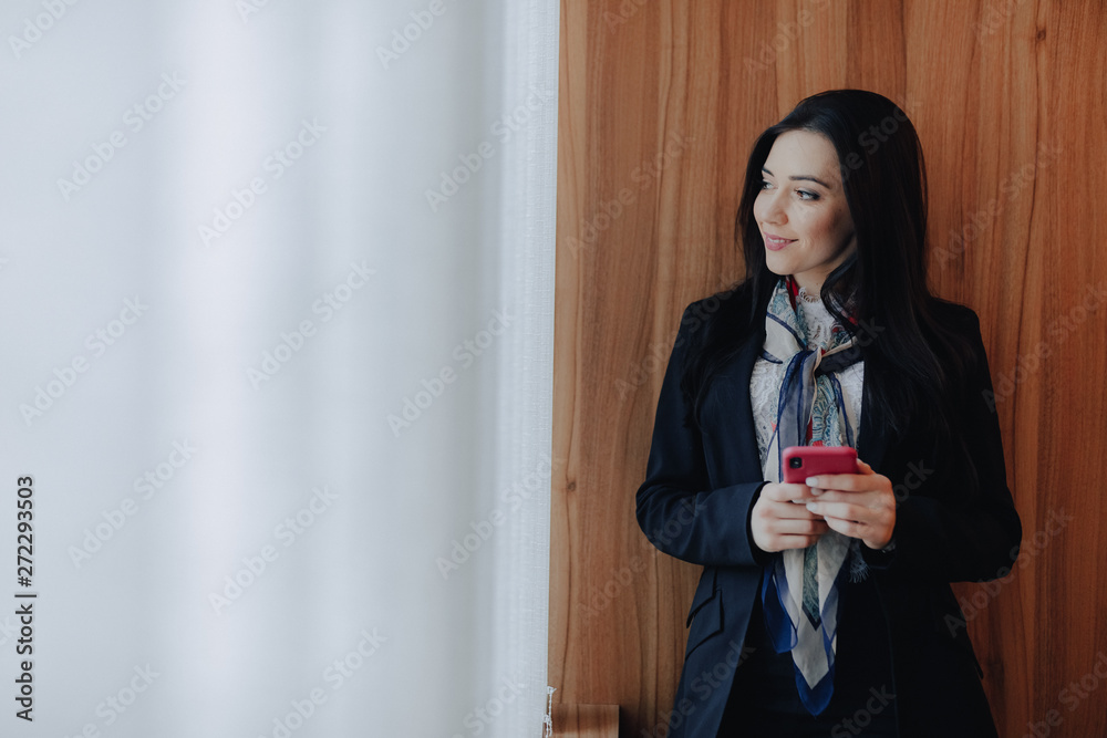young emotional attractive girl in business-style clothes at a window with a telephone in a modern office or auditorium young emotional attractive girl in business-style clothes at a window with a telephone in a modern office or auditorium