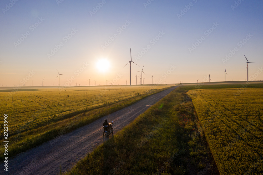 Biker is standing on a road between wheat and wind turbines in the sunrise light. Empty road to ride a motorbike. Eolian generators in a beautiful wheat field. Wind turbine. Wheat Field Agriculture Biker is standing on a road between wheat and wind turbines in the sunrise light. Empty road to ride a motorbike. Eolian generators in a beautiful wheat field. Wind turbine. Wheat Field Agriculture