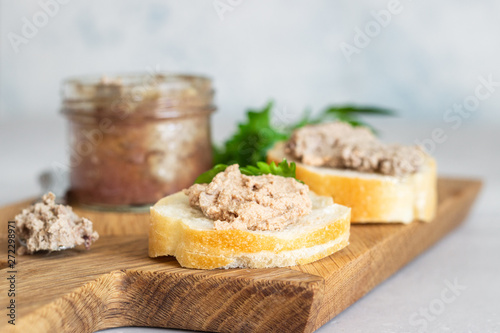 Liver pate in a glass jar with fresh bread and parsley on a wooden cutting board. Light grey stone background.