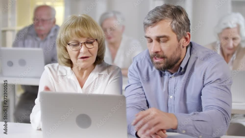 Tracking shot of middle-aged male technology teacher helping elderly woman using laptops in computer class Tracking shot of middle-aged male technology teacher helping elderly woman using laptops in computer class