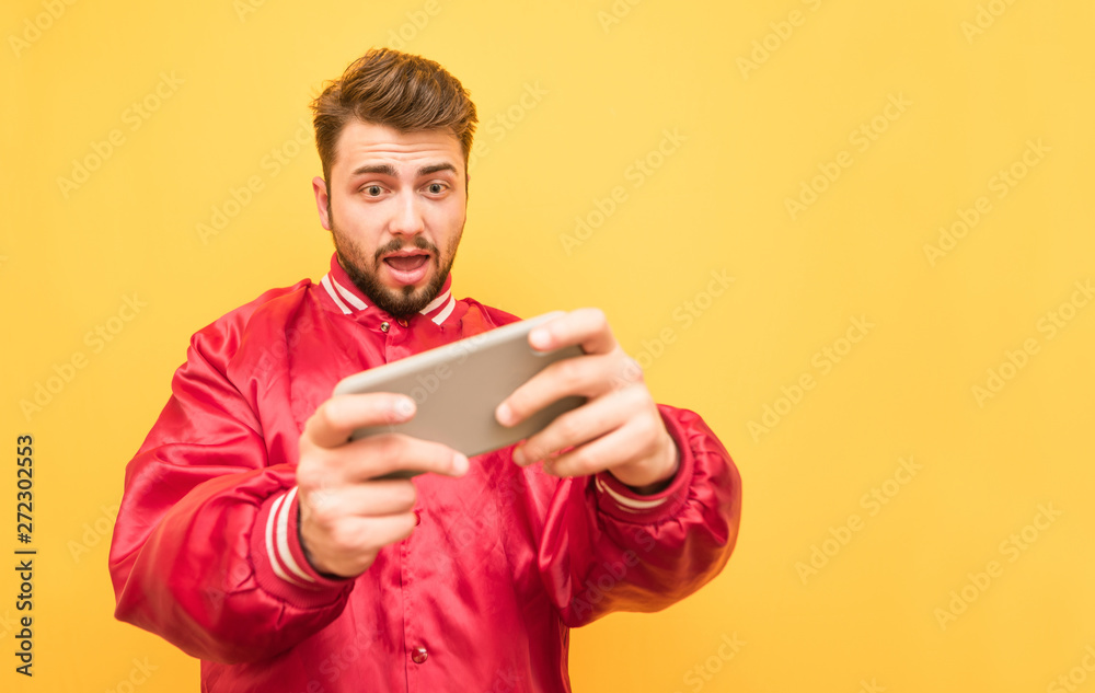 Portrait of an adult playing mobile games on a smartphone, focused looking at the screen, standing on a yellow background in a red jacket. Adult gamer plays mobile games, isolated. Portrait of an adult playing mobile games on a smartphone, focused looking at the screen, standing on a yellow background in a red jacket. Adult gamer plays mobile games, isolated.