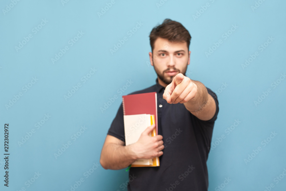 Male student with books in his hands stands on a blue background and shows the index finger in the camera wearing a black shirt and beard. Isolated. Male student with books in his hands stands on a blue background and shows the index finger in the camera wearing a black shirt and beard. Isolated.
