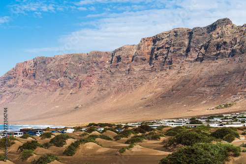 Sand dunes shaped like waves and covered in green vegetation at the iconic Famara Beach. Rugged volcanic sheer cliffs in Lanzarote, Canary, Spain.