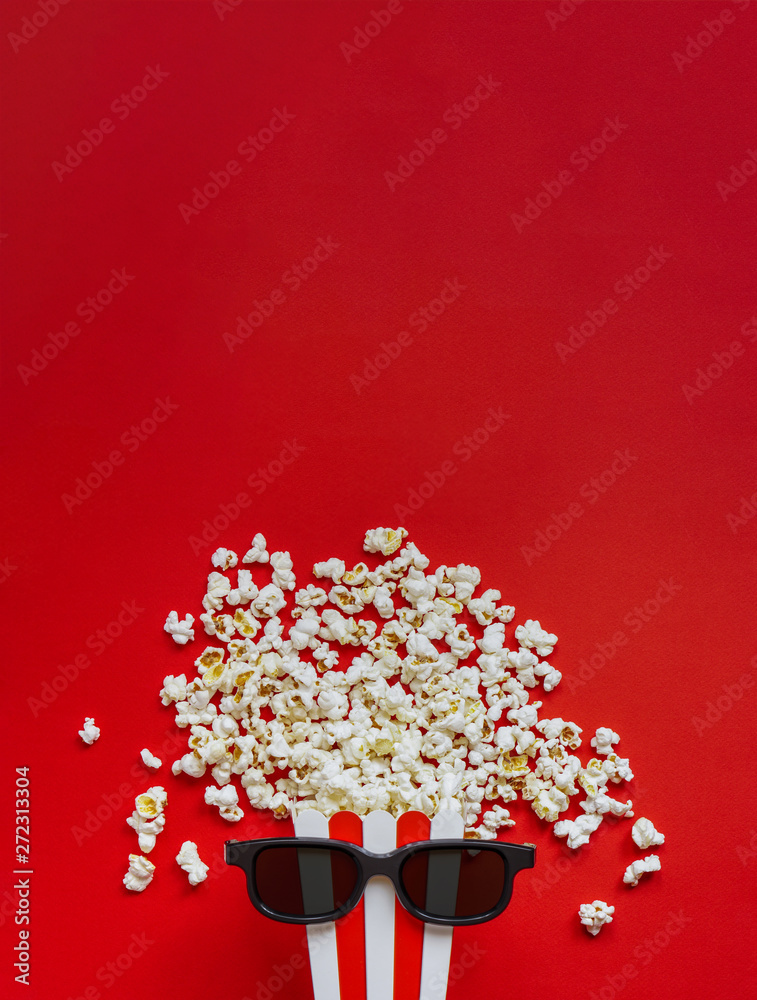 Popcorn in a red and white cardboard box with glasses for a 3D movie on the red background. Popcorn in red striped bucket on red back. Flat lay concept. Popcorn in a red and white cardboard box with glasses for a 3D movie on the red background. Popcorn in red striped bucket on red back. Flat lay concept.