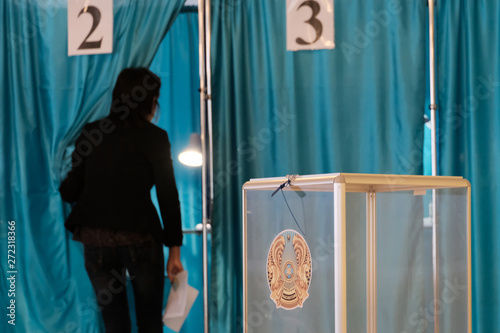 Kazakhstan, Qazaqstan, June 9, 2019, Elections, Voting room. A woman walks into an individual booth with numbers two. Transparent box with the emblem of Kazakhstan for counting votes. Blue background. © romsvetnik