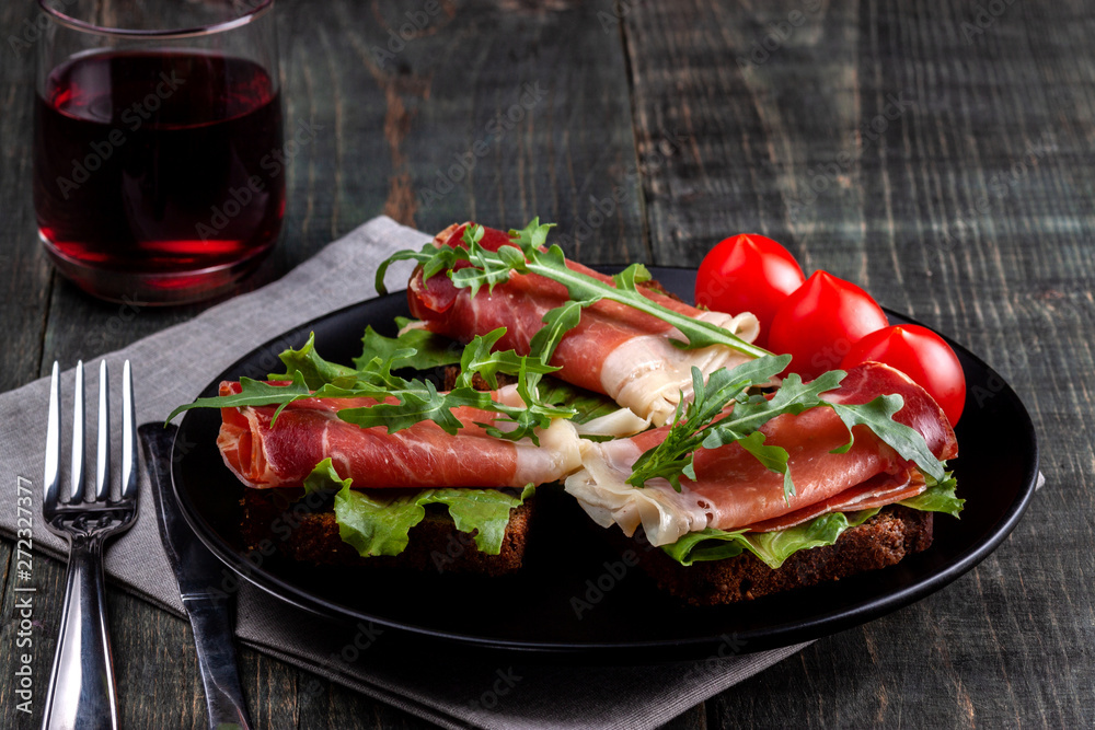 On the table there is a dish with jamon sandwiches, tomato, greens and a glass of red wine On the table there is a dish with jamon sandwiches, tomato, greens and a glass of red wine
