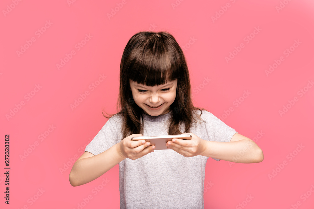 Image of emotional young pretty cute little girl posing isolated over pink studio wall background play games by mobile smart sell phone.