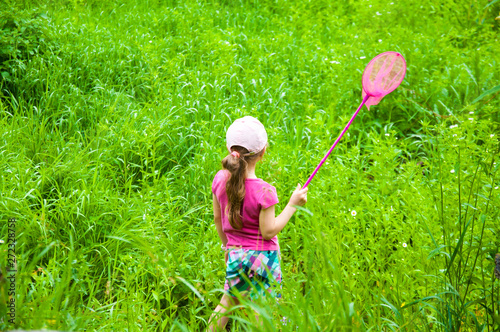 A little girl in a pink T-shirt stands with a net in her hands. Catching butterflies. On a green meadow in summer. Green grass.