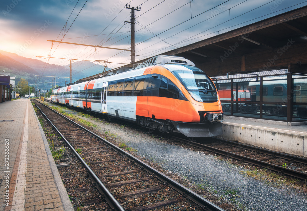 High speed train on the railway station in mountains at sunset in summer. Orange modern commuter train on the railway platform. Industrial landscape with railroad. Passenger transportation. Intercity