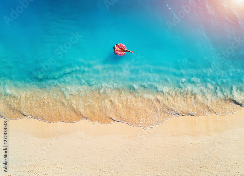 Aerial view of a young woman swimming with the donut swim ring in the clear blue sea with waves at sunrise in summer. Tropical aerial landscape with girl, azure water, sandy beach. Top view. Travel © den-belitsky