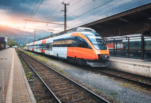 Obraz Wysoki prędkość pociąg na stacji kolejowej w górach przy zmierzchem w lecie. Pomarańczowy nowoczesny pociąg podmiejski na peronie kolejowym. Krajobraz przemysłowy z kolei. Transport pasażerski. Międzymiastowy