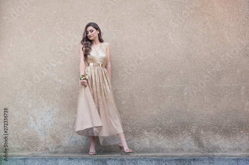 Fototapeta Beautiful teen girl in glamorous golden dress standing by the wall