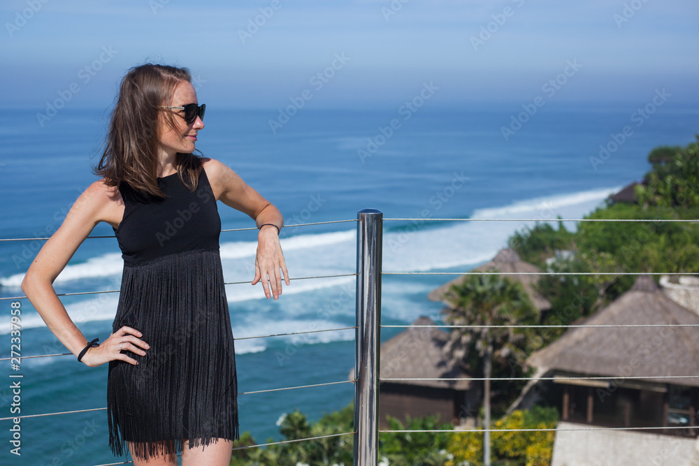 A beautiful miniature slender young woman in a black dress and sunglasses is standing on a terrace leaning on a railing. Blue sky and blue ocean in the background. sunny day