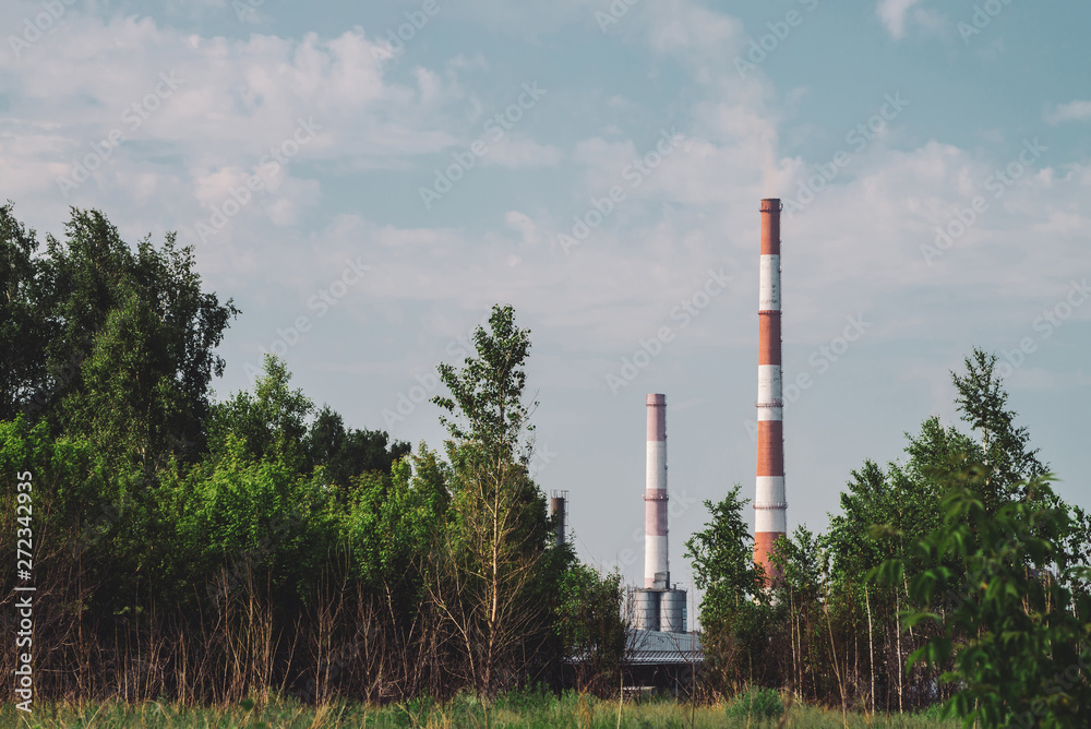 Giant smoke stack in industrial area behind trees. Industrial structure with big pipe of brown brick under blue sky. Technology and nature. Pollution of environment. Vegetation in industry background. Giant smoke stack in industrial area behind trees. Industrial structure with big pipe of brown brick under blue sky. Technology and nature. Pollution of environment. Vegetation in industry background.