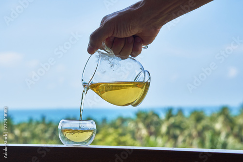  Close male hand with glass tea kettle pouring tea into glass Chinese traditional cup against the backdrop on the sea, blue sky and trees