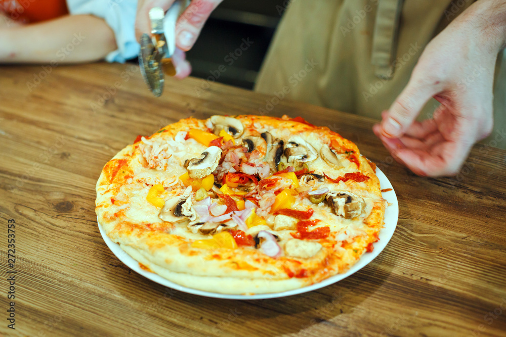 Cutting freshly cooked homemade pizza that lies on a white plate on a wooden tabl