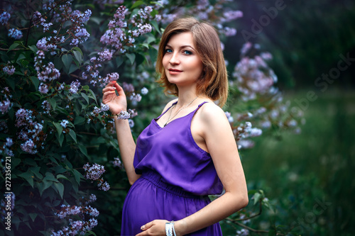 A charming beautiful young pregnant woman in purple violet dress in a blooming lilac garden looks at camera with tenderness. Beautiful nature bokeh in the background © Anastassiya 
