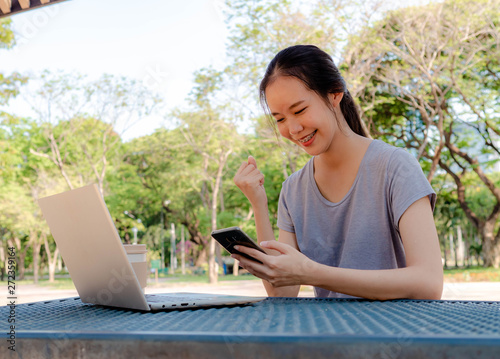 Happy young girl with a phone in her hands working on a laptop at the table in the park. The concept of shopping online in the Internet, freelance working concept.