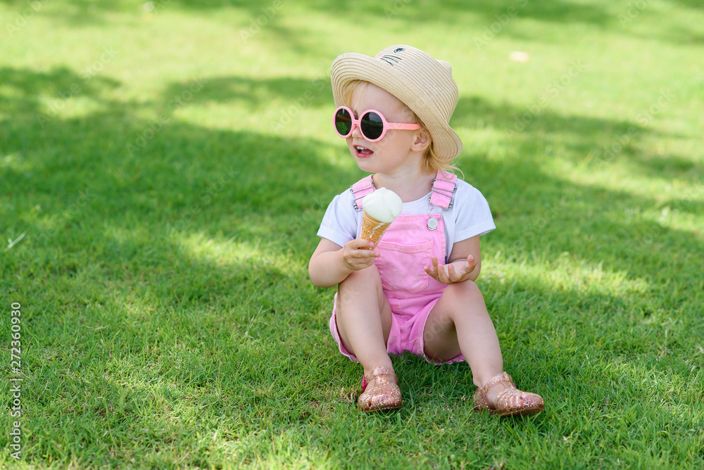 Happy toddler girl in summer hat and pink sunglasses eats with pleasure tasty ice cream while sitting on a green grass on hot sunny day. Happy toddler girl in summer hat and pink sunglasses eats with pleasure tasty ice cream while sitting on a green grass on hot sunny day.