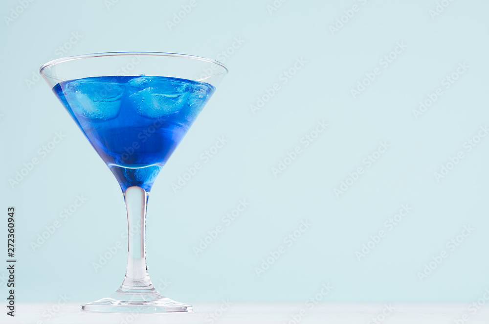 Summer cold blue lagoon drink with ice cubes in elegant martini glass on pastel mint background.