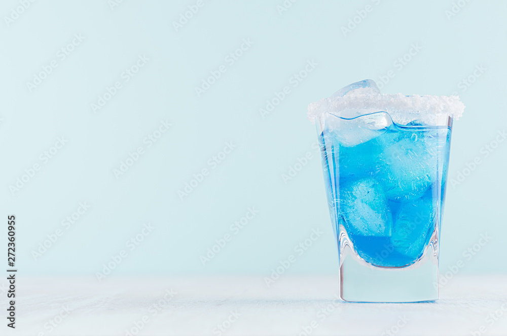 Tropical fresh alcohol cocktail with blue curacao liqueur, ice cube, salt rim in frozen shot glass on soft light white, pastel green background. Tropical fresh alcohol cocktail with blue curacao liqueur, ice cube, salt rim in frozen shot glass on soft light white, pastel green background.