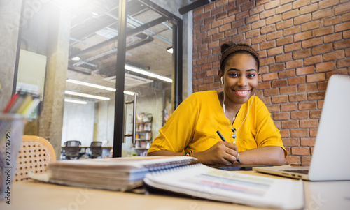 Portrait of beautiful smile black girl read books listen to earphones in library campus or modern office. Young creative casual business woman working on desk with computer photo