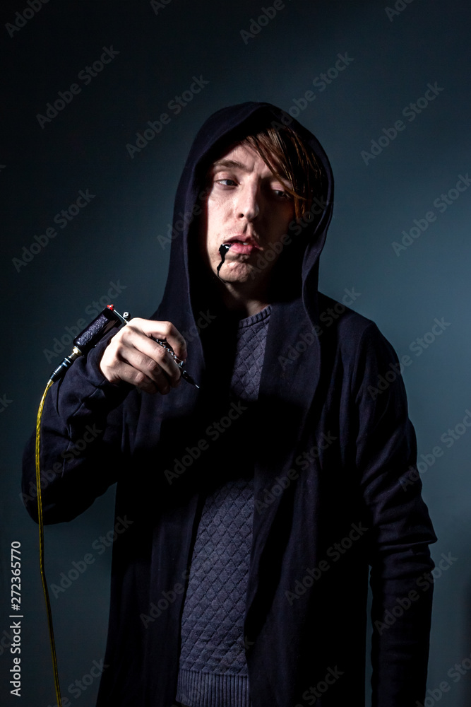 unusual freak young nasty guy in black hood and red bangs stand and look with paint face and tattoo machine in studio with deep shadows and cold isolated background. concept of death, despair and fear